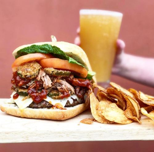 A stacked burger with pulled pork, pickles, lettuce, and tomato, served with chips and a glass of beer.