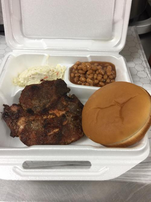 A takeout container with a grilled meat portion, coleslaw, baked beans, and a soft sandwich bun.