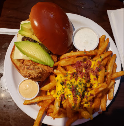 A plate with a chicken sandwich topped with avocado and cheese, served with loaded cheese fries and dipping sauces.