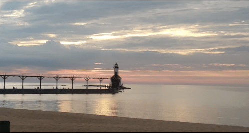 A serene lighthouse at sunset, reflecting on calm waters with a cloudy sky and sandy beach in the foreground.
