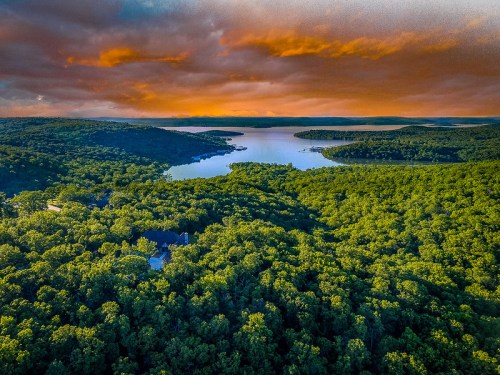 Aerial view of lush green forests surrounding a tranquil lake under a vibrant orange and purple sunset sky.