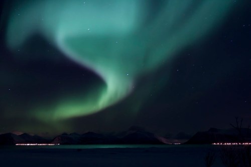 A stunning display of the Northern Lights illuminates the night sky over a snowy landscape and distant mountains.