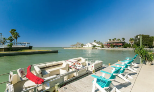 A serene waterfront view with a boat dock, turquoise chairs, and palm trees under a clear blue sky.