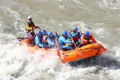 A group of six people in blue and orange life jackets paddling a bright orange raft through whitewater rapids.