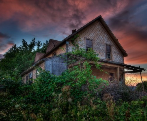 Abandoned house overgrown with vines, set against a dramatic sunset sky with colorful clouds.
