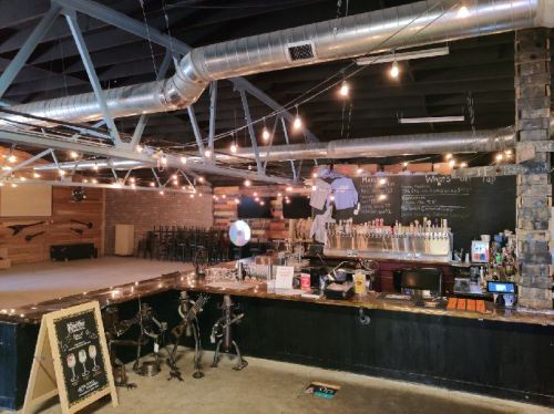 Interior of a bar with a wooden counter, beer taps, string lights, and a stage area in the background.
