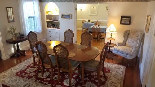 A cozy dining room with a large wooden table, chairs, and a patterned rug, leading to a well-lit kitchen area.