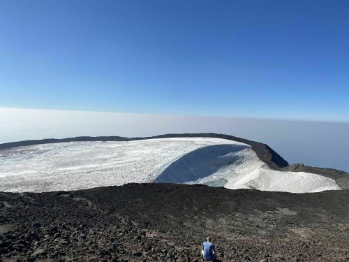 A person sits on rocky terrain overlooking a vast glacier and clear blue sky on a volcanic landscape.
