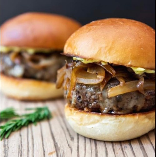 Two juicy burgers with caramelized onions and mustard on soft, toasted buns, placed on a wooden surface.