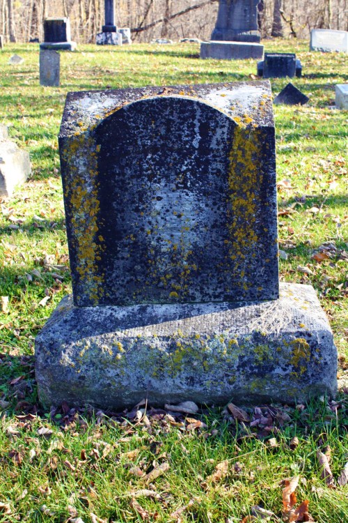 A weathered gravestone covered in moss and lichen, set in a grassy cemetery with other stones in the background.