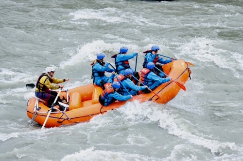 A group of six people in blue gear navigating a turbulent river in an orange raft, with one person steering.