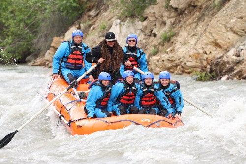 A group of seven people in blue and red rafting gear paddles an orange raft on a river surrounded by rocky terrain.