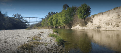 A serene riverbank scene with a bridge in the background, surrounded by trees and rocky shores under a clear sky.