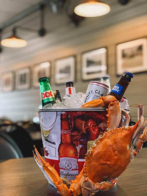 A bucket filled with ice, assorted beer bottles, and a large crab on a wooden table in a cozy restaurant setting.