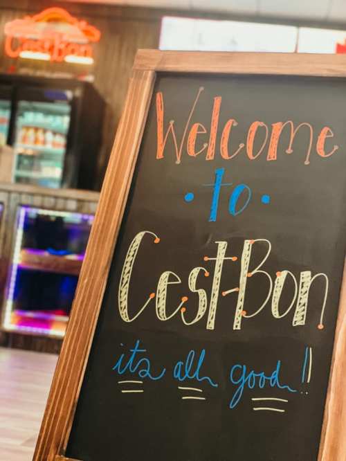 A wooden sign reads "Welcome to Cest Bon! It's all good!" in colorful chalk lettering.