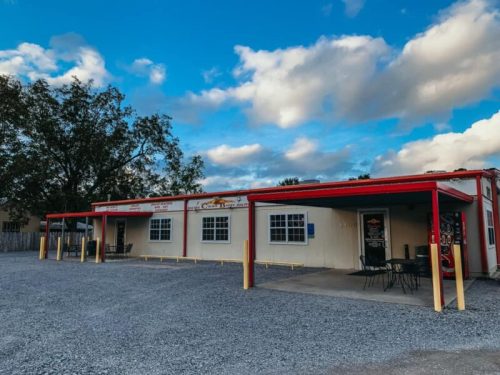 A small, single-story building with a red roof and outdoor seating, set against a cloudy sky. Gravel parking area in front.