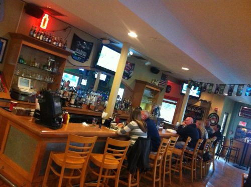 A bar scene with patrons seated at the counter, a well-stocked bar, and sports on TV in a casual setting.