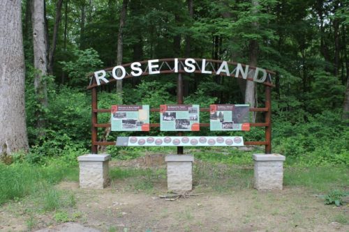 Sign at Rose Island with historical information, surrounded by lush green trees in a forested area.