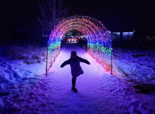 A child joyfully spins under a colorful light arch on a snowy path at night.