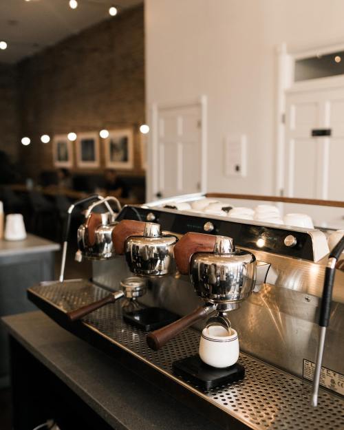 A close-up of a coffee machine with shiny metal portafilters and a small milk pitcher on a countertop in a café.