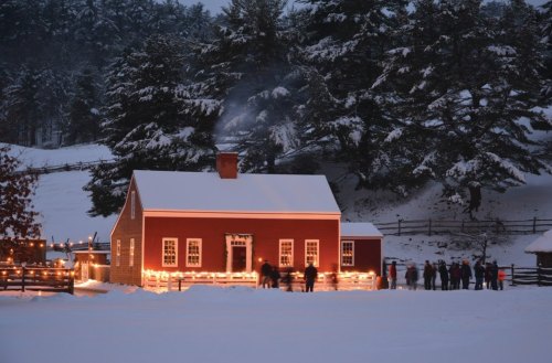 A cozy red house with snow-covered roof, warmly lit, surrounded by trees and people enjoying a winter evening.