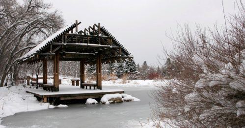 A wooden pavilion on a frozen pond, surrounded by snow-covered trees and shrubs in a winter landscape.