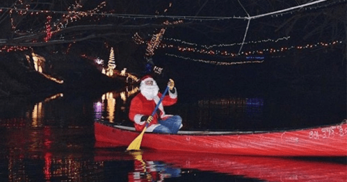 Santa in a red canoe paddles through a festive, illuminated waterway decorated with holiday lights.