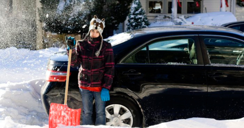 A person in a raccoon hat and gloves stands with a shovel in a snowy driveway beside a parked car.