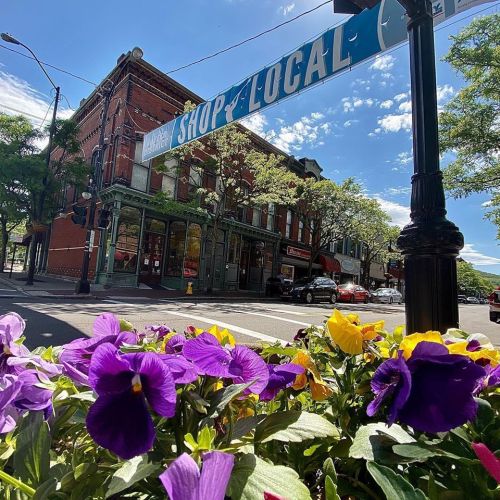 A vibrant street scene featuring a "Shop Local" banner above colorful flowers and historic buildings under a blue sky.