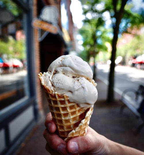 A hand holding a waffle cone filled with two scoops of ice cream, with a tree-lined street in the background.