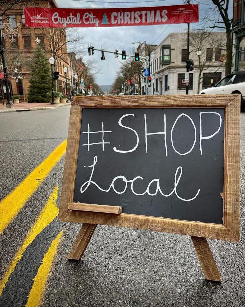 A chalkboard sign on a street reads "#SHOP Local," with a festive banner overhead in a quiet town setting.