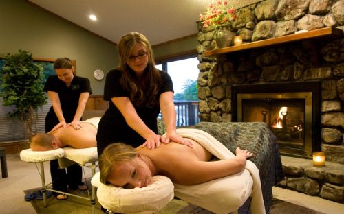 Two people receiving massages in a cozy room with a stone fireplace, while a therapist works on one of them.