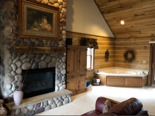 Cozy cabin interior featuring a stone fireplace, wooden walls, a large tub, and rustic decor.