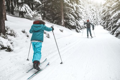 A child skiing on a snowy trail, with a person skiing in the background among snow-covered trees.
