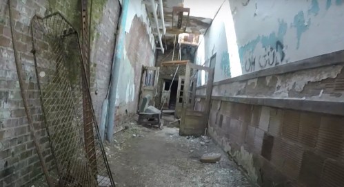 An abandoned hallway with crumbling walls, debris, and a broken door leading to a dark space.