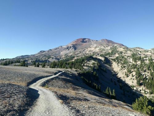 A winding dirt path leads through a rocky landscape towards a mountain under a clear blue sky.