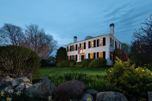 A charming white house with black shutters, surrounded by greenery and flowers, illuminated at dusk.
