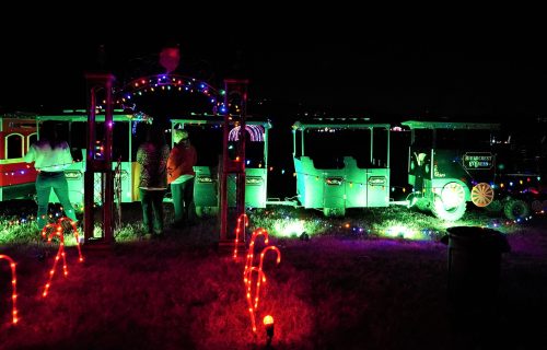 Colorful holiday lights illuminate a train display at night, with people interacting near the festive decorations.