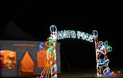 A brightly lit "North Pole" sign with festive decorations, next to a tent glowing with warm light at night.