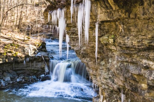 A serene waterfall cascades over rocks, surrounded by icicles and a forest in early spring.