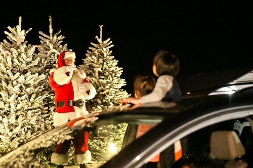 Santa Claus stands near decorated trees, while two children watch from a car, enjoying the festive atmosphere.