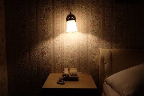 A bedside table with a vintage phone and a lamp casting light on floral wallpaper in a dimly lit room.