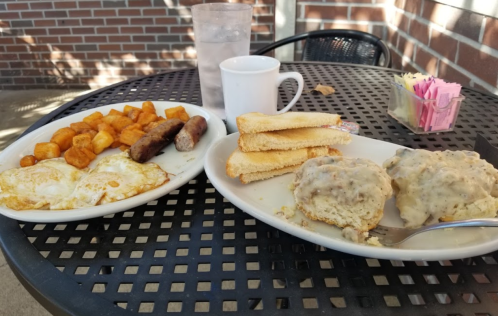A breakfast plate with eggs, sausage, biscuits with gravy, and toast, alongside a glass of water and a coffee cup.