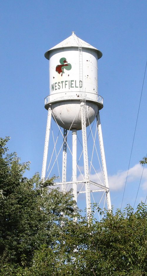 A white water tower with "WESTFIELD" painted on it, surrounded by green trees against a blue sky.