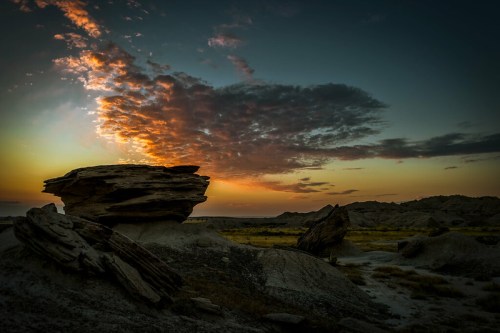 A dramatic sunset over a rocky landscape, with clouds illuminated in vibrant colors against a darkening sky.