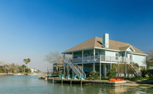 A light blue house by the water with a dock, surrounded by palm trees and clear blue skies.