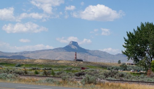 A distant mountain rises against a blue sky with clouds, near a grassy area and a small building with a chimney.