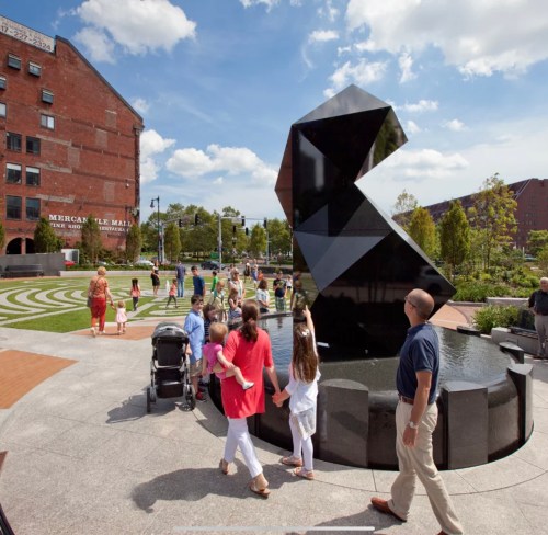 A modern black sculpture in a park with people walking, children playing, and buildings in the background under a blue sky.