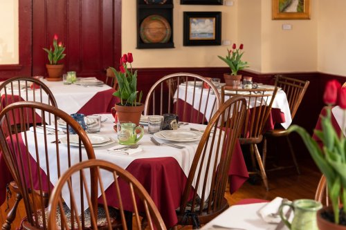 Cozy restaurant interior with wooden chairs, white tablecloths, and red accents, featuring potted tulips on tables.