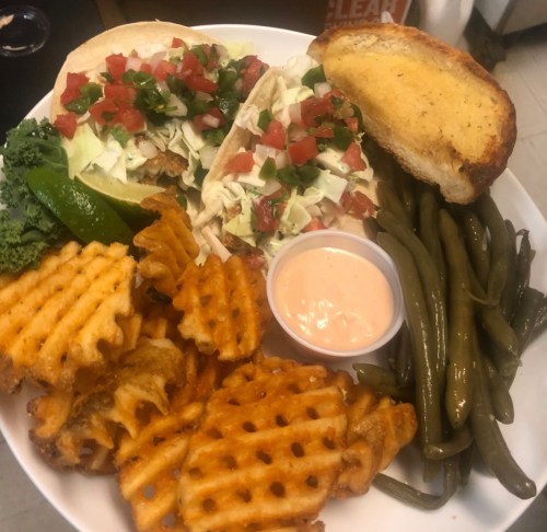 A plate with two tacos topped with lettuce and tomatoes, waffle fries, green beans, and a slice of garlic bread.